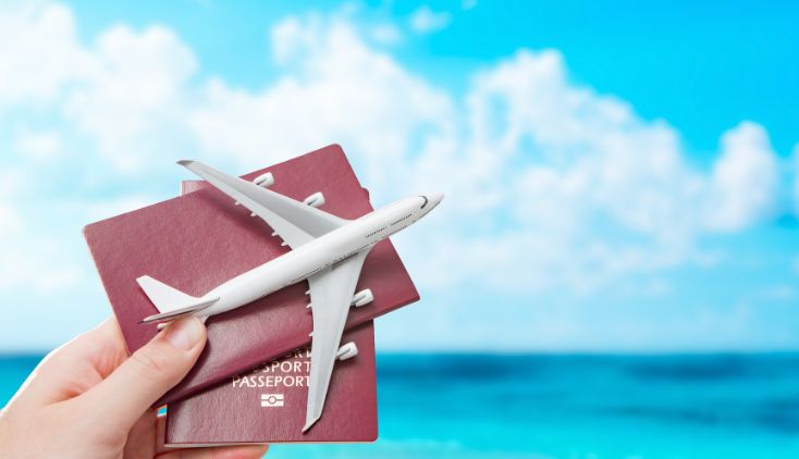 A hand holding two passports and a toy airplane in front of a beach.