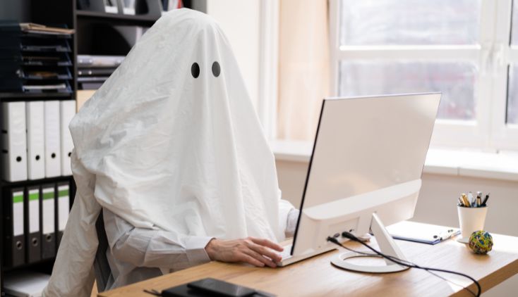 A person sitting at a desk with a computer, wearing a ghost costume.