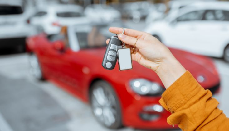 A hand holding a set of car keys in front of a red car.
