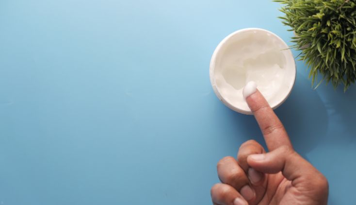 A finger with face cream on it, above a container of the cream on a blue background.