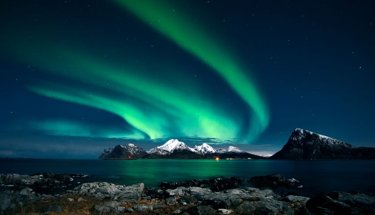 A photo of the Northern Lights over water and snow capped mountains.